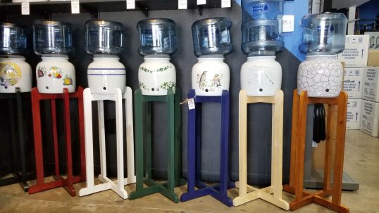 An assortment of water bottle stands for your reverse osmosis water. Stands shown are red, white, green, blue, neutral wood and stained wood